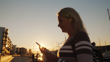 A-Woman-In-Headphones-Uses-A-Smartphone-On-The-Pier-On-The-Background-Of-Private-Yachts-At-Sunset-Th