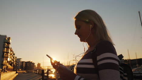 uma mulher em fones de ouvido usa um smartphone no cais no fundo de iates privados ao pôr do sol th