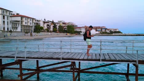 Athletic-young-male-walking-across-a-scenic-coastal-wooden-pier-boardwalk-wearing-backpack-and-sunglasses