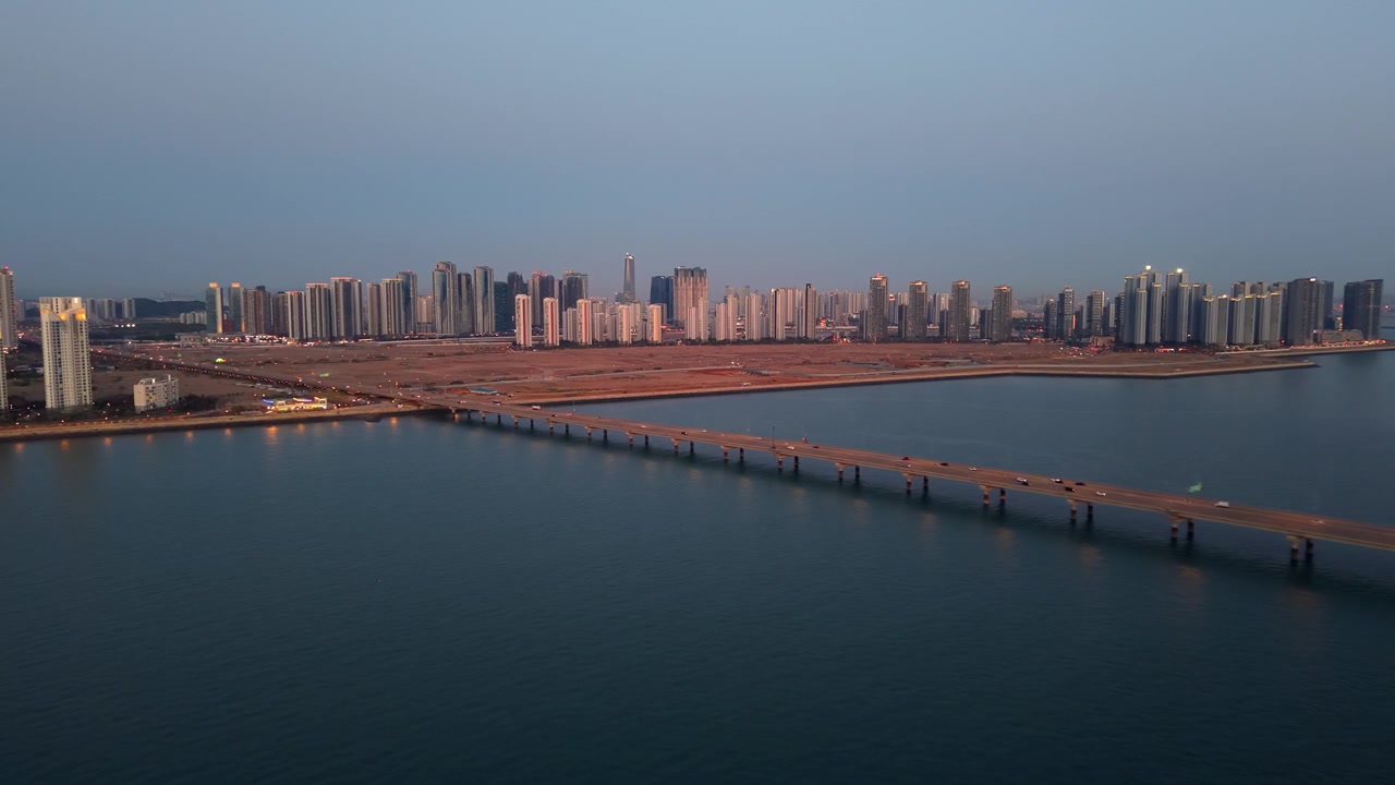 Aerial view moves forward across bay toward busy causeway and dense Songdo skyscraper skyline during blue hour dusk