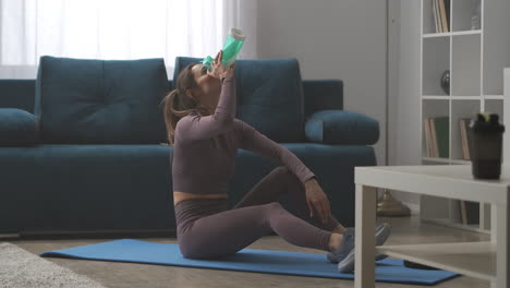 mujer deportiva se relaja después del entrenamiento en casa sentada en el suelo en la sala de estar bebiendo agua de botellas de plástico deportivas deporte y estilo de vida saludable