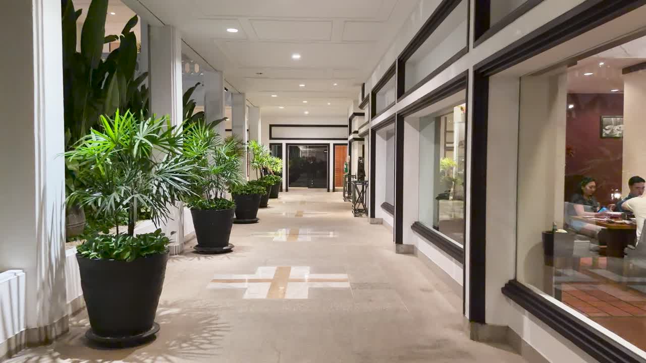 Steady camera glides down well-lit hotel corridor with lush plants, glass walls, evening ambiance