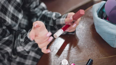 vista parcial de primer plano de una mujer sosteniendo diferentes artículos de maquillaje, incluidos lápiz labial y brillo labial, mientras contempla su elección, una bolsa de maquilaje con esponjas y polvo compacto es visible en la mesa