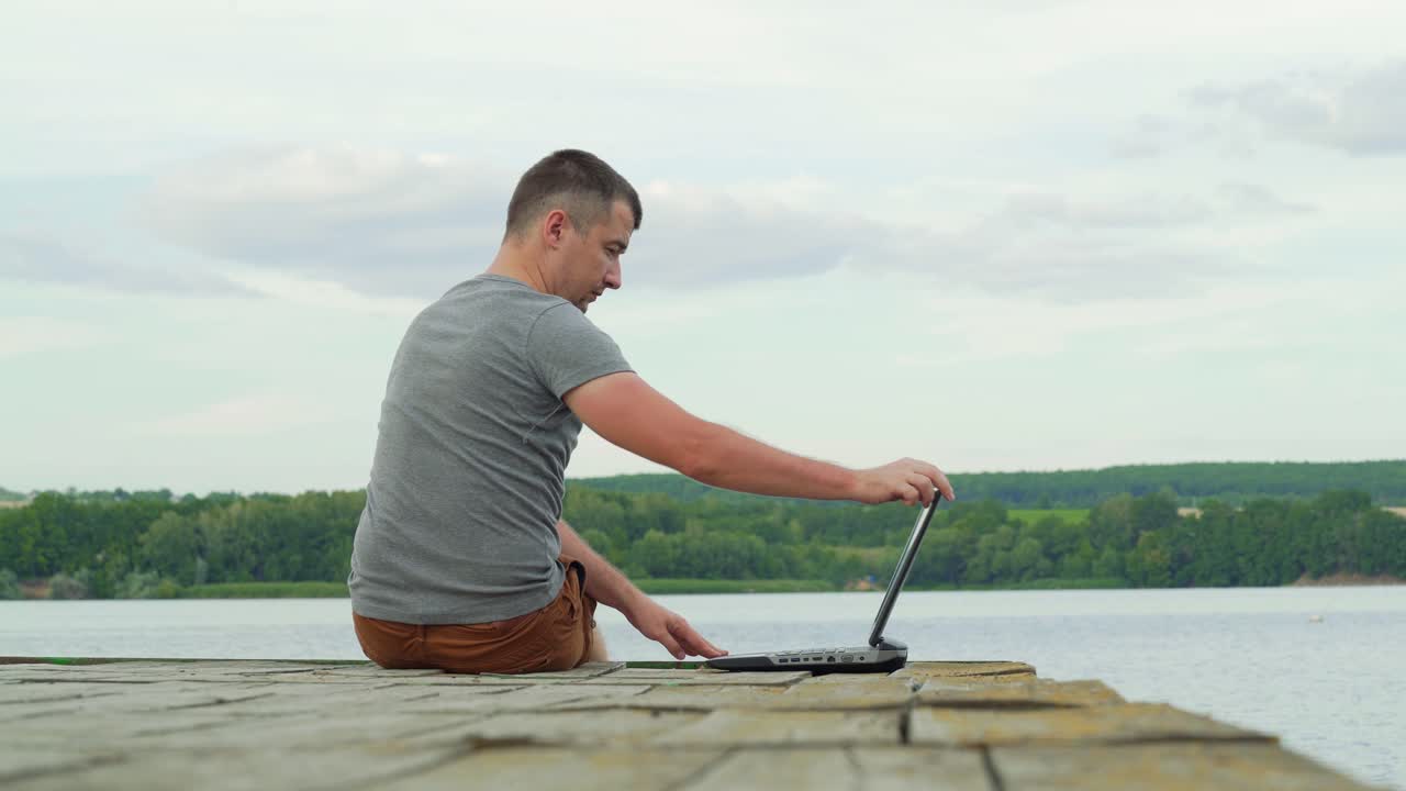 Young male sitting at the edge of the bridge and opens a laptop and started working near the river. Man in a grey t-shirt working on nature with modern gadget