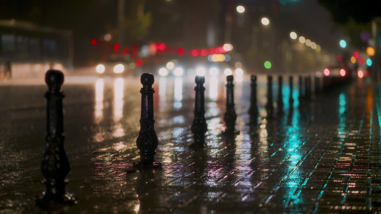 Rain falls steadily on city streets, creating mirrored reflections on the pavement. Streetlights and vehicle headlights illuminate the scene, adding vibrant colors to the rainy atmosphere