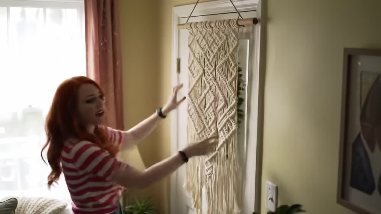A Creative Woman Shows Her Craftsmanship by Hanging a Beautifully Made Macramé Wall Hanging in a Brightly Lit Living Room, Adding a Personal Touch to the Decor