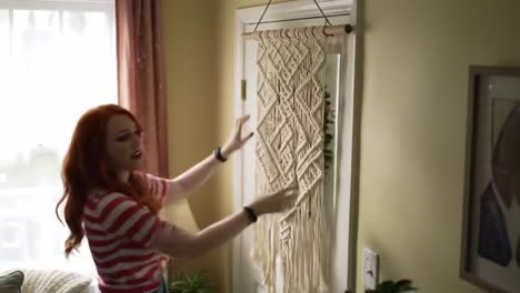 a creative woman shows her craftsmanship by hanging a beautifully made macramé wall hanging in a brightly lit living room, adding a personal touch to the decor