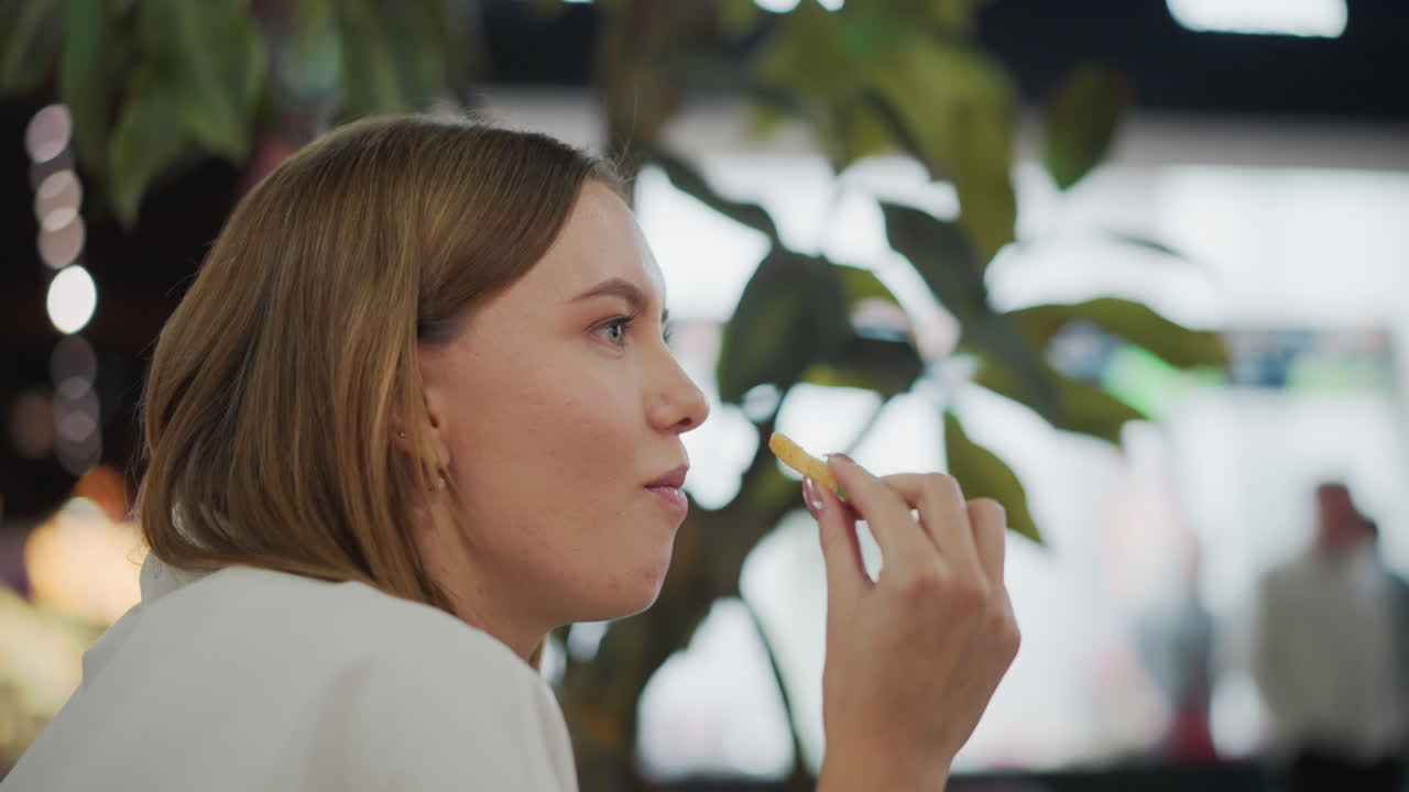 fotografía en primer plano de una mujer con uñas pulidas comiendo papas fritas crujientes en un entorno de comedor casual, con un fondo suave y borroso con iluminación cálida y personas entrando en el centro comercial