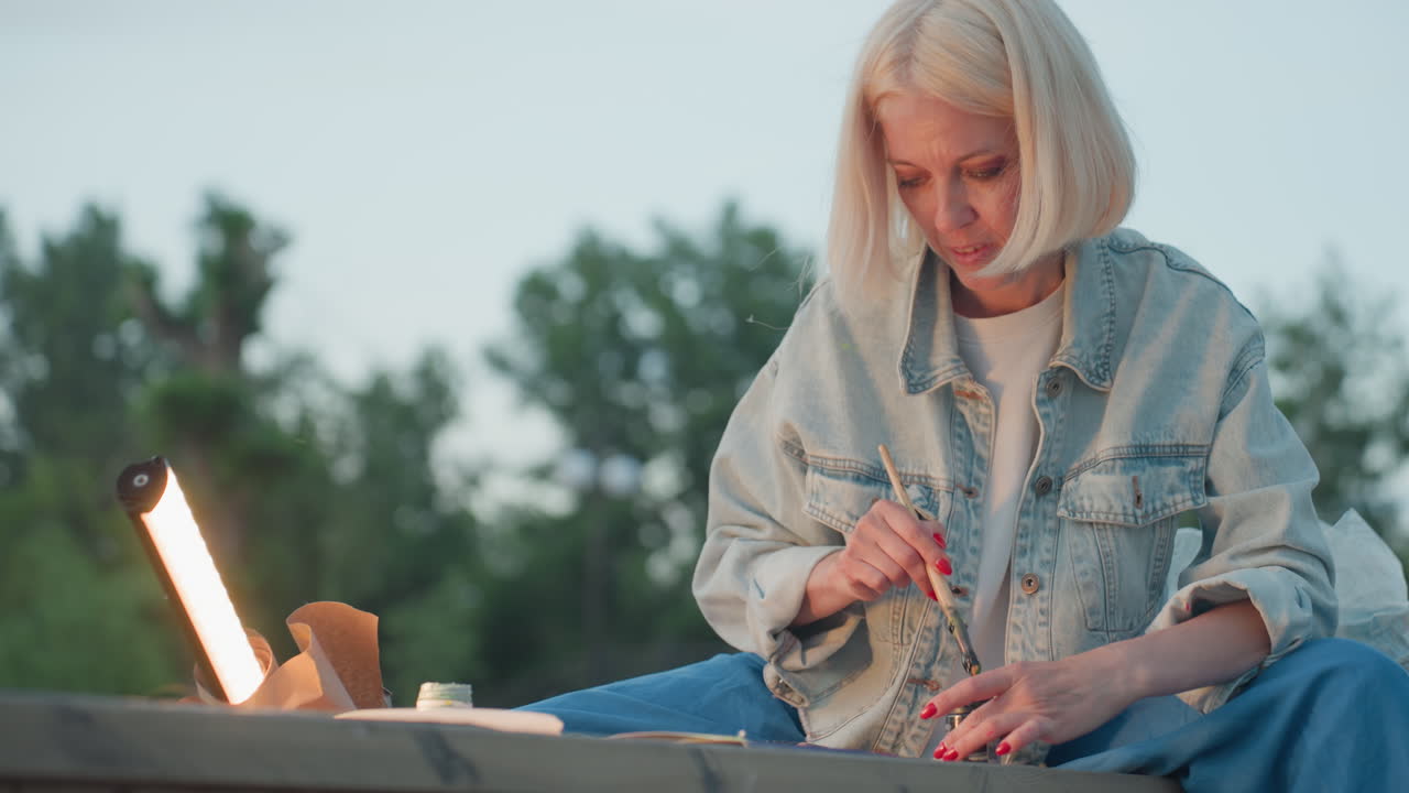 close up fine artist seated on bench outdoors painting detailed strokes under lamp shine with focused expression while children play nearby framed by twilight park trees and jars of paint