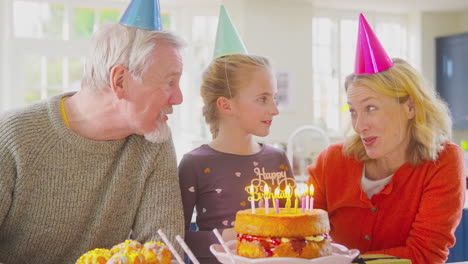 Abuelos-Con-Nieta-Celebrando-Juntos-Soplando-Velas-En-El-Pastel-De-Cumpleaños-En-Casa