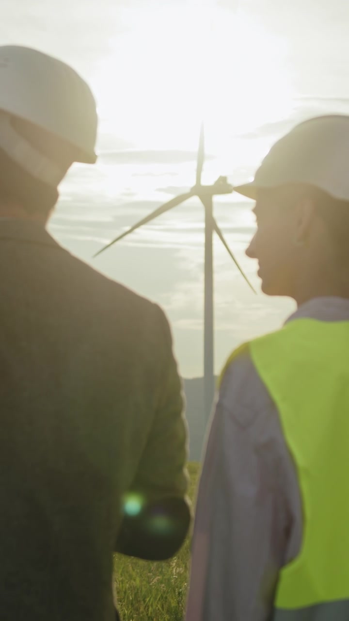 Engineers in hard hats and safety vests observe wind turbines in a grassy field at sunset discussing