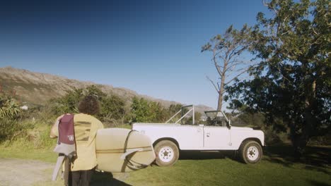 follow shot of surfer walking towards land rover and loading his surfboard on to the back with wetsuit over the shoulder