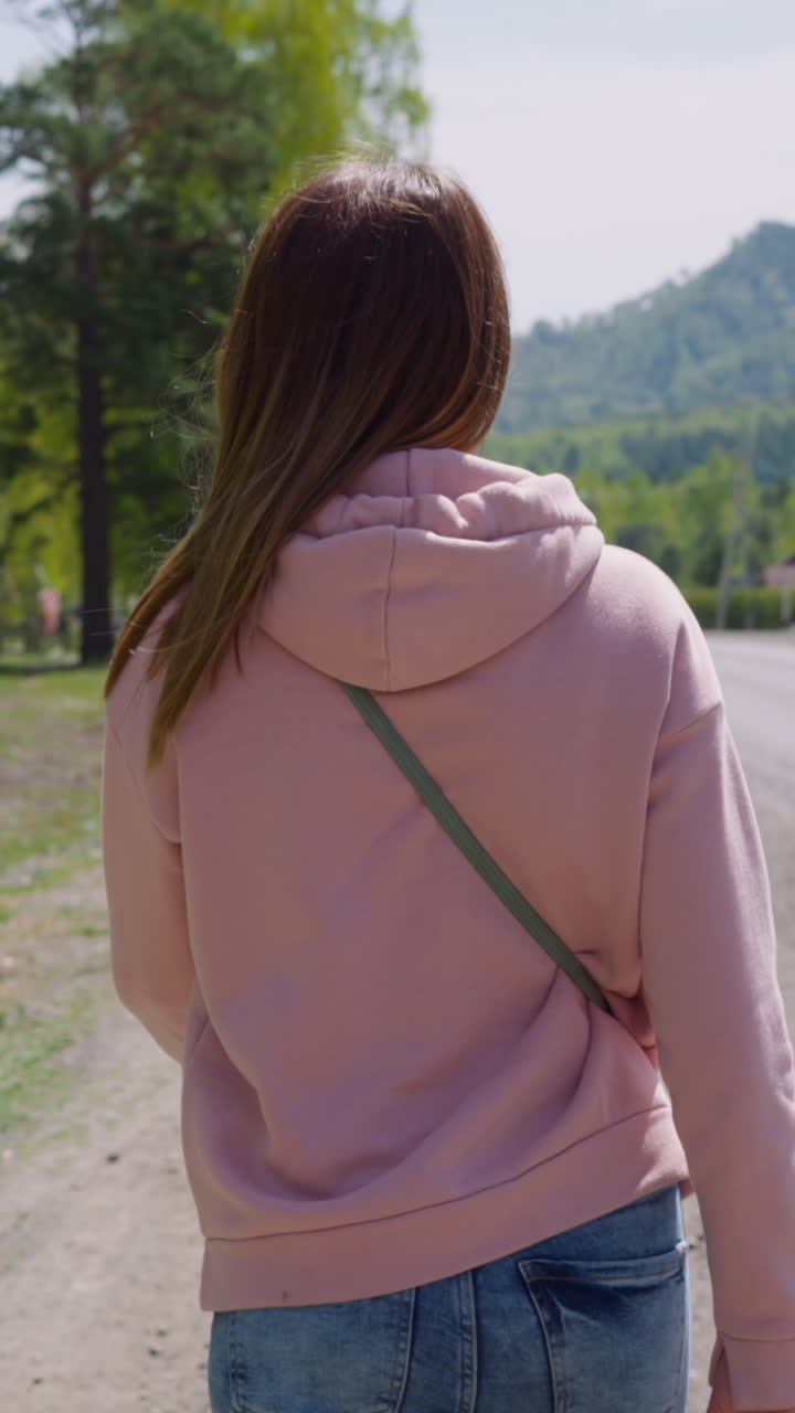 Long haired woman in pink hoodie walks along rural road at eco resort backside view. Tourist spends vacation at highland. Wild nature conservation