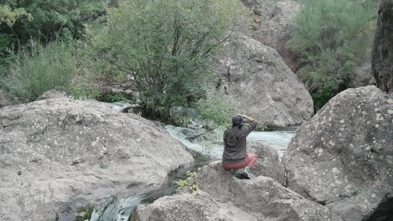 Brunette girl facing away, capturing photos of the Rio Mundo waterfall, in a rugged landscape with greenery