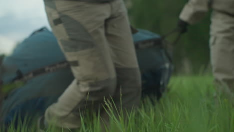 close up of two people dragging gear bag through tall grass toward paramotor trailer under soft sky, focus on steady posture and teamwork emphasising adventure prep in outdoor field