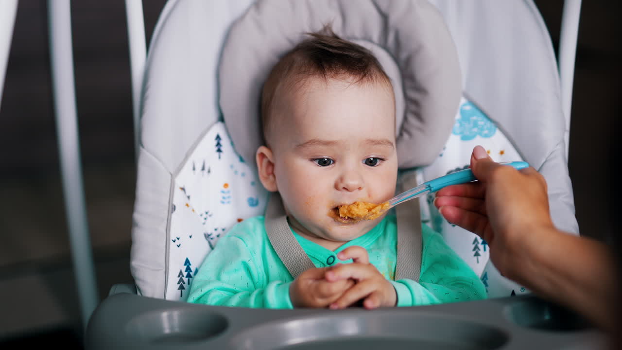 Mother giving food on spoon to her son. Baby opens mouth and doesn't like it making face and waving hands.