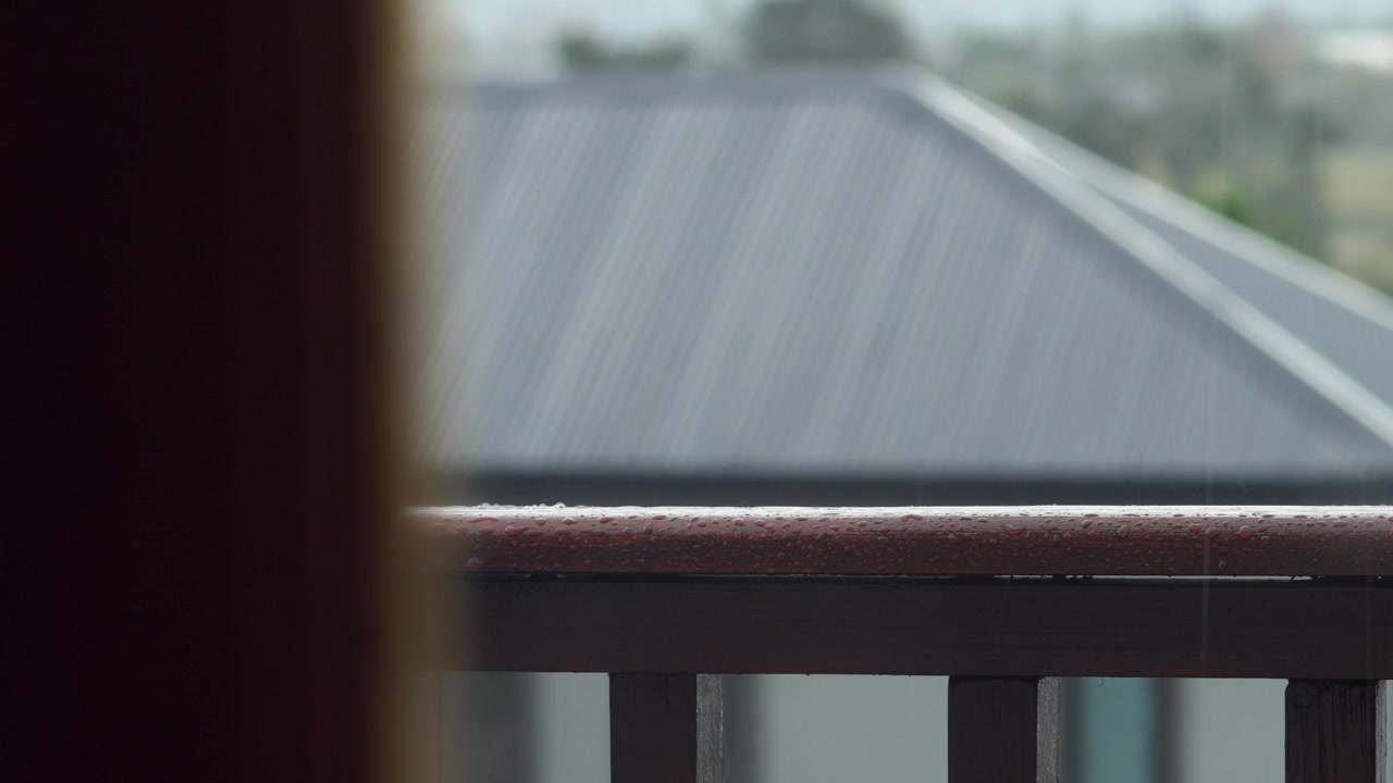 Heavy raindrops bouncing off a wet brown wooden balcony handrail. A dark corrugated metal roof of a neighboring house sits blurred in the background. Static close up view.
