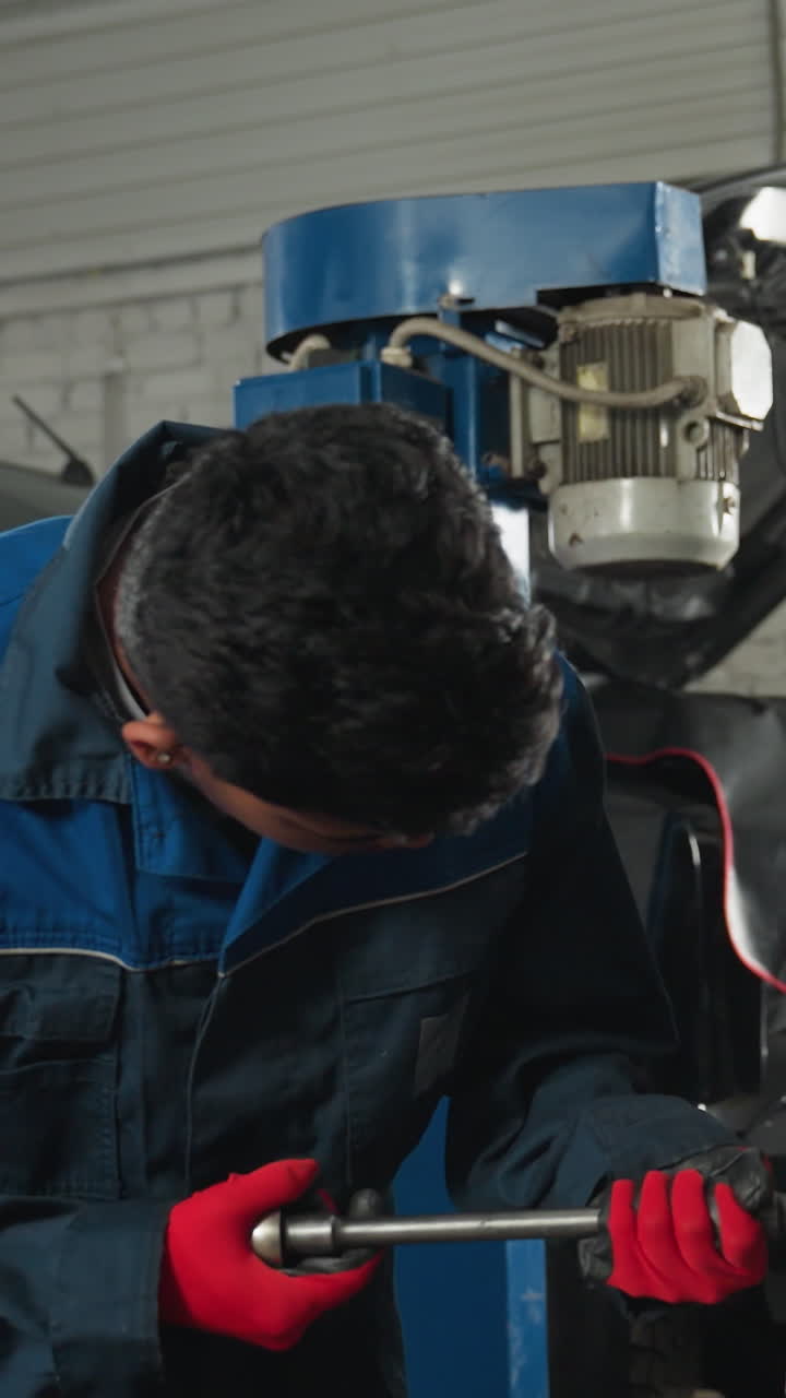 ingenieros mecánicos en un taller; uno trabajando atentamente en un motor con una llave inglesa mientras su colega usa su teléfono, iluminado por una luz brillante en el fondo