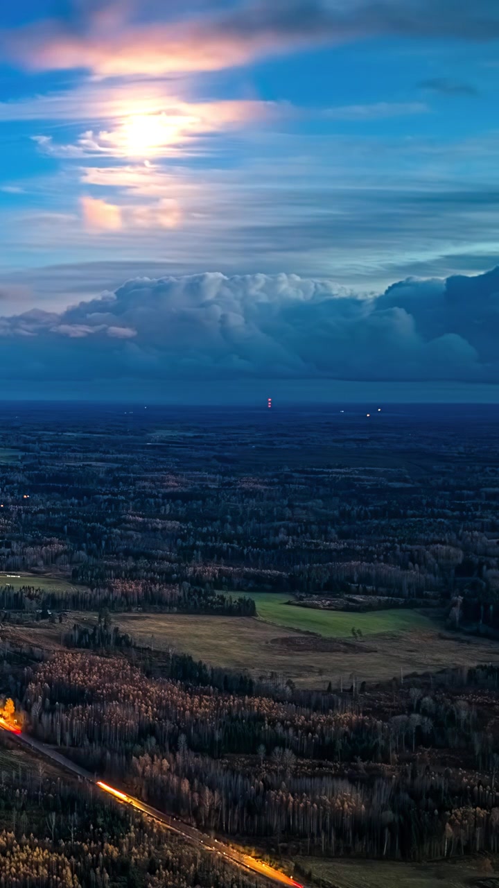 Timelapse verticale a lunga esposizione aerea cattura la luna luminosa con alone lunare, nuvole drammatiche e fluttuanti e scie luminose di auto su strada di campagna sopra una vasta foresta autunnale lettone all'alba