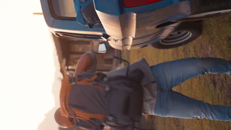 Vertical-Video-Portrait-Of-Man-Unloading-Backpacks-From-Pick-Up-Truck-On-Road-Trip-In-Countryside