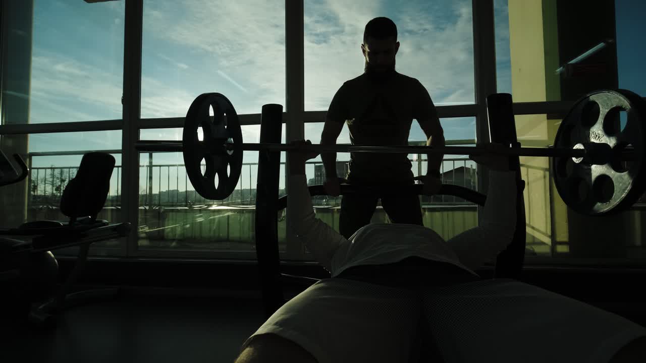 트레이너는 바벨, 벤치 프레스를 들어 올리는 것을 감독합니다. 남자는 체육관의 벤치에 누워있는 바의 도움으로 가과 팔의 근육을 흔들고 있습니다.