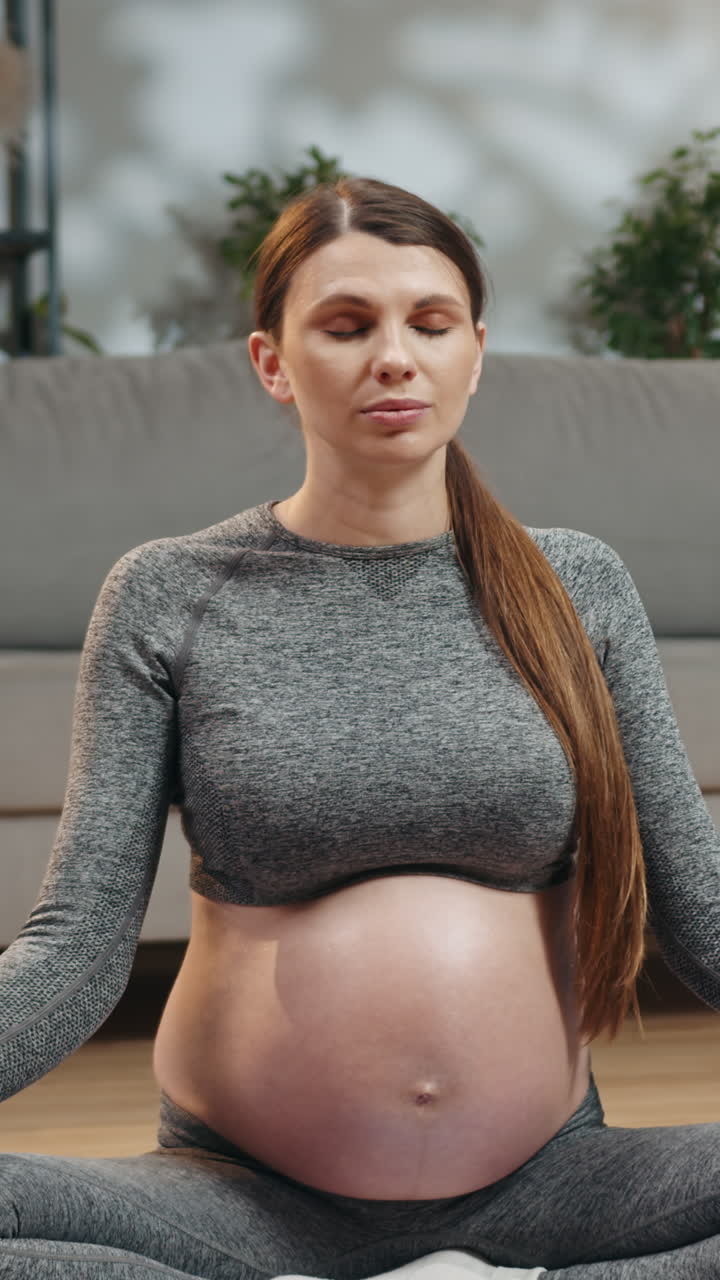 Pregnant woman in sportswear meditating with eyes closed on yoga mat in living room practicing