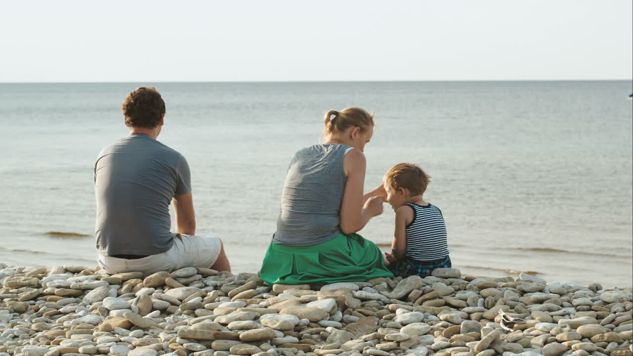 familia de tres sentados en la playa de guijarros junto al agua