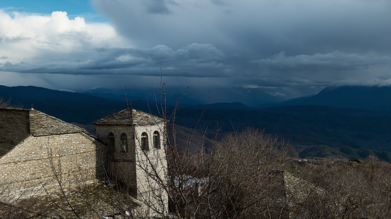 A historic stone building stands against a backdrop of dramatic clouds and mountains in Zagori. Ideal for travel and cultural documentaries.