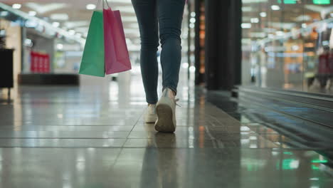 leg view of a woman in jeans and sneakers carrying mint and pink shopping bags while walking through a shopping mall with glossy tiled floors and colorful light reflections