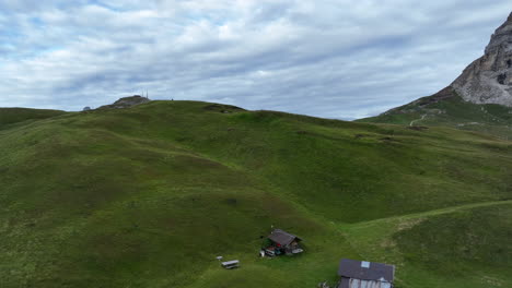 Green-hilly-meadow-and-small-home-with-Dolomites-in-background,-aerial-view