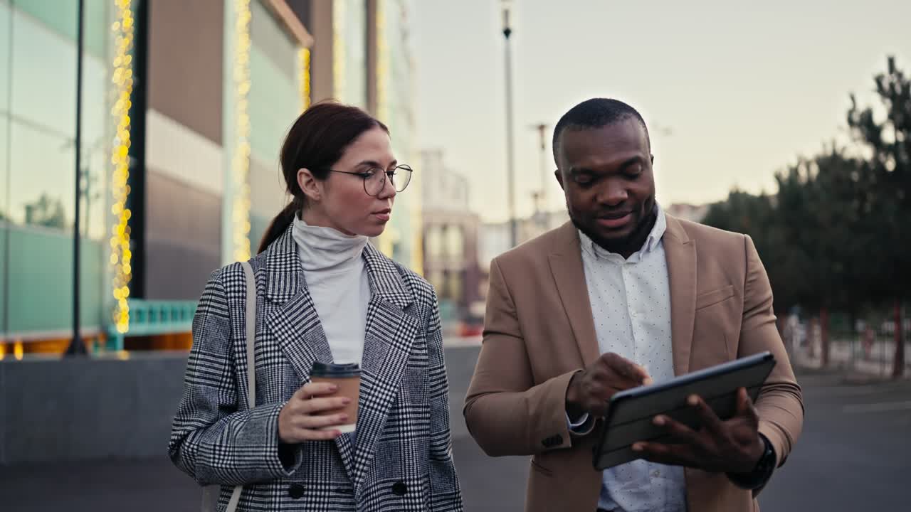 A confident male manager with Black skin, a short haircut and a beard in a brown suit walks and talks with his colleague a businesswoman a brunette in round glasses and a gray coat, and looks at something on a Black tablet during his business meeting in the city