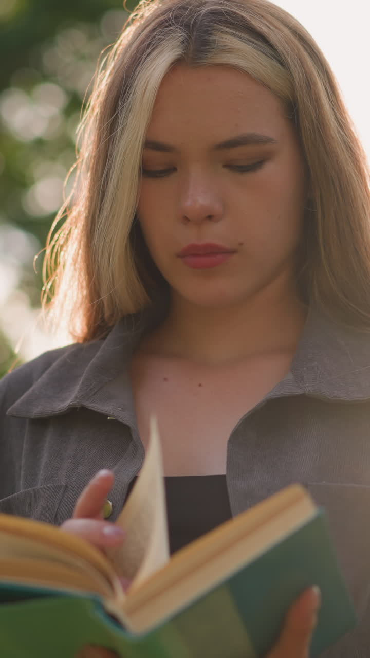 dama con ropa gris de pie al aire libre, cabeza ligeramente inclinada, hojeando las páginas de un libro con la luz del sol reflejándose suavemente en el fondo características borrosos árboles y edificios