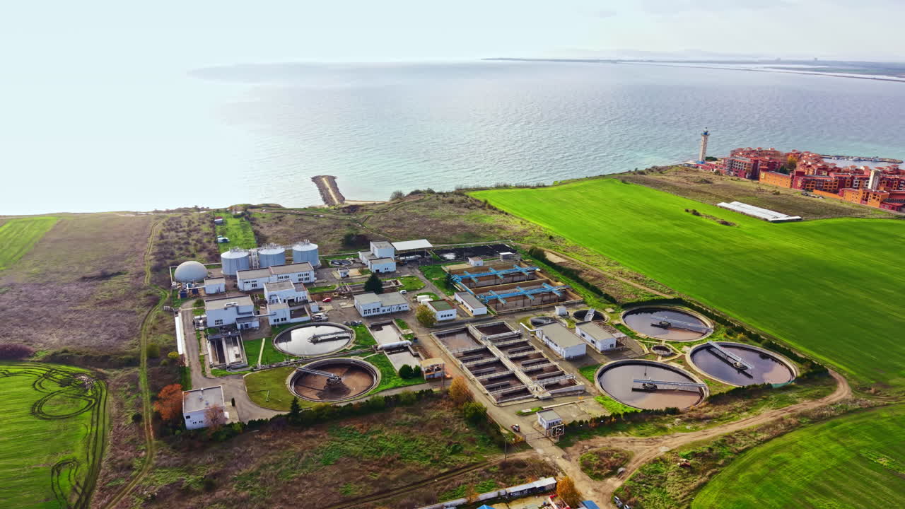 Aerial view captures a wastewater treatment plant beside a serene shoreline and lush green fields during a bright day