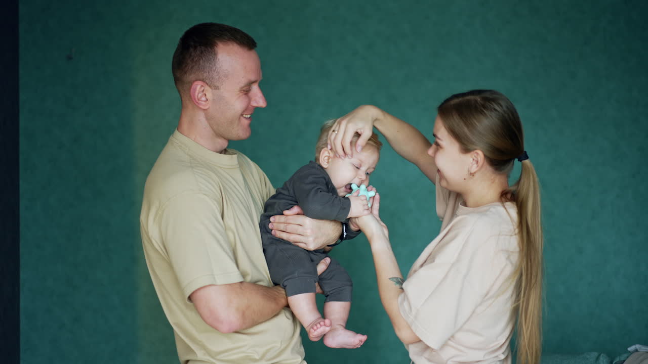 Caucasian man holding a baby with blond hair. Mother gives a rubber toy to her child and touches his hair.