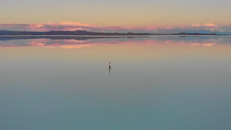 Luftaufnahme-Einer-Einsamen-Figur,-Die-In-Der-Abenddämmerung-Auf-Dem-Spiegelbild-Der-Größten-Salzwüste-Der-Welt-In-Den-Salzwüsten-Von-Uyuni,-Bolivien,-Steht