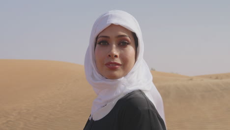 ritratto di una bella donna musulmana in hijab bianco e abito nero tradizionale in piedi in un deserto ventoso e sorridente alla telecamera 1