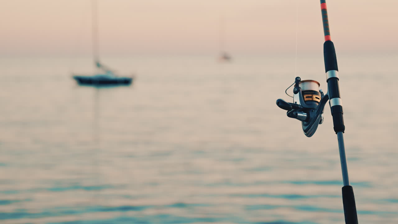 Primo piano di una canna da pesca e un mulinello posizionati vicino al mare, con una barca sfocata sullo sfondo
