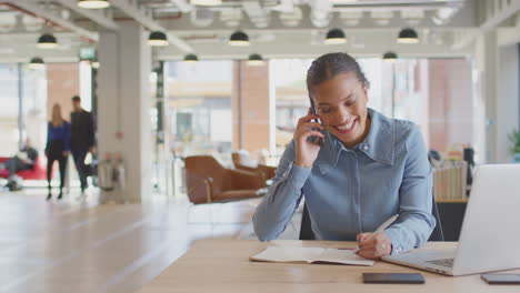 Businesswoman-Sitting-At-Desk-On-Phone-Call-In-Modern-Open-Plan-Office-With-Colleagues-In-Background