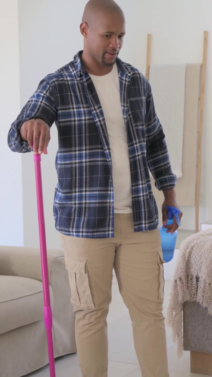 Vertical video: Entering room, man in plaid spraying floor, cleaning with pink mop and blue bottle