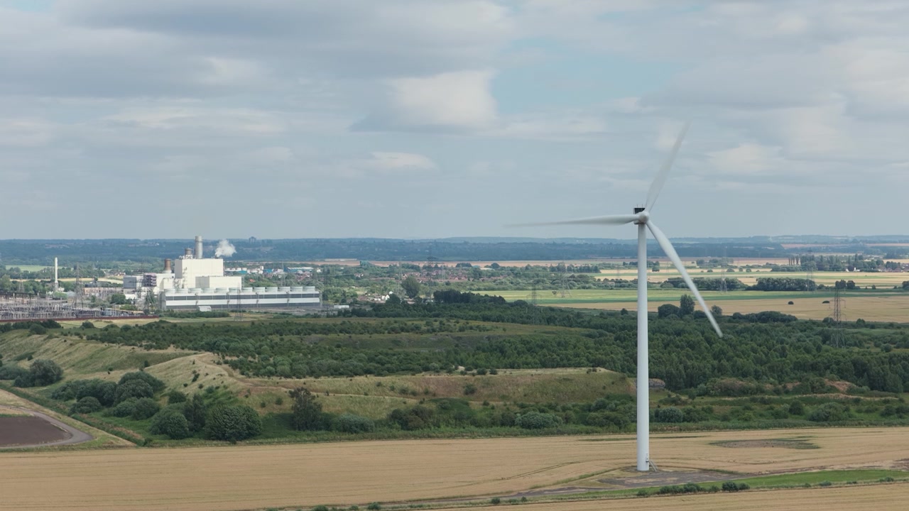A large wind turbine efficiently spins its blades in an expansive rural field. In the distance, an industrial power plant stands against a bright, partly cloudy daytime sky.
