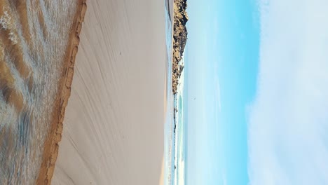 Zambujeira-do-Mar-over-the-sea-shore-with-ocean-waves,-cliffs-and-sand-dunes-covered-by-green-vegetation-red-leaves-of-sour-fig,-sunny-day,-clear-blue-sky