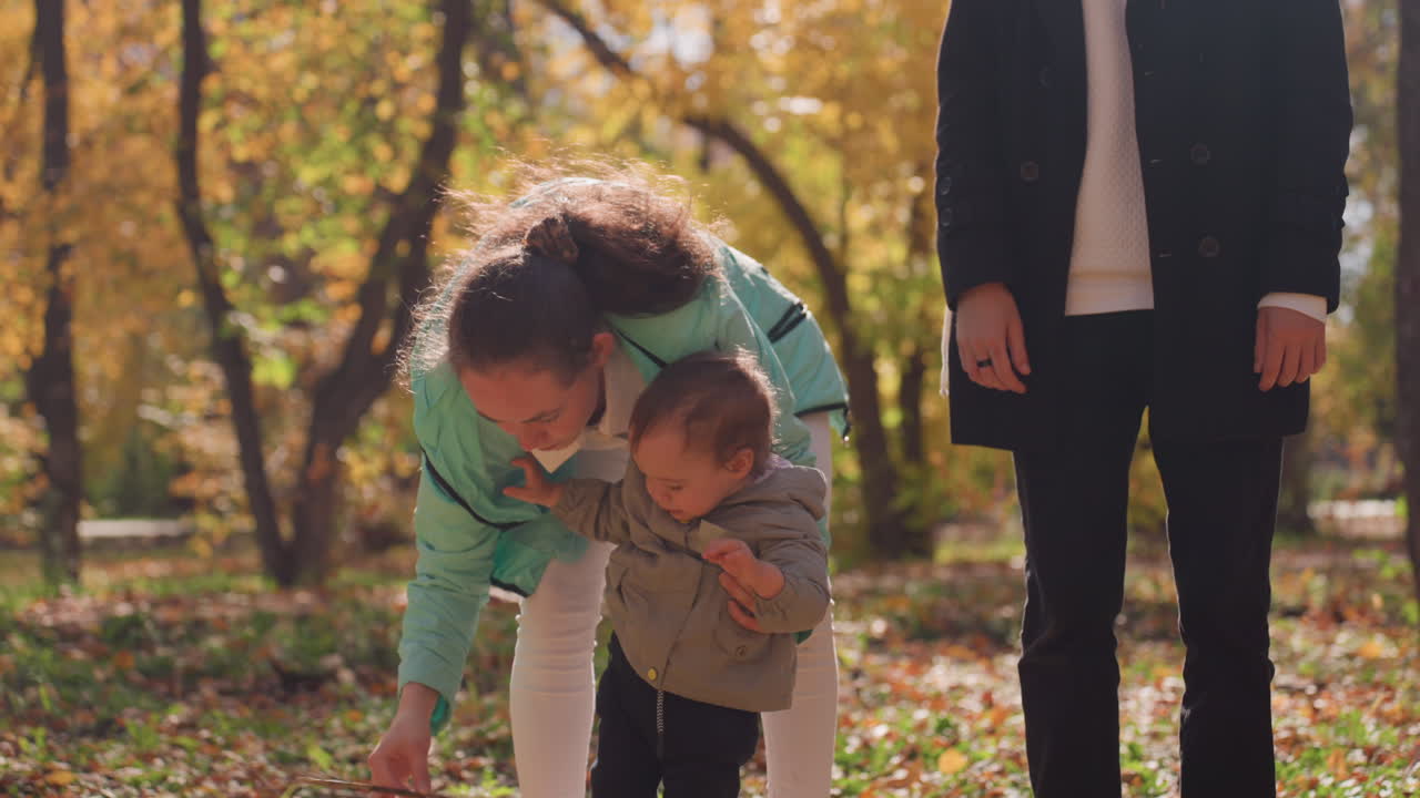 Madre bianca nel parco autunnale che insegna al bambino a camminare, guida delicata e mani ferme, il padre osserva nelle vicinanze, foglie cadute sotto i piedi, incoraggiamento paziente, un momento familiare significativo.