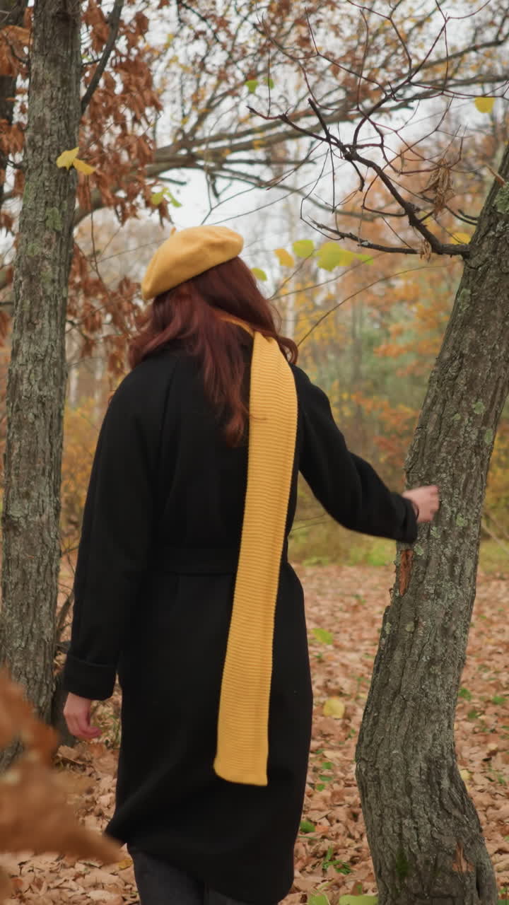 Bakfra av en kvinne i en svart kåpe og gult skjerf som går gjennom en skog, berører tørre blader og trebark, fordypet i naturen.