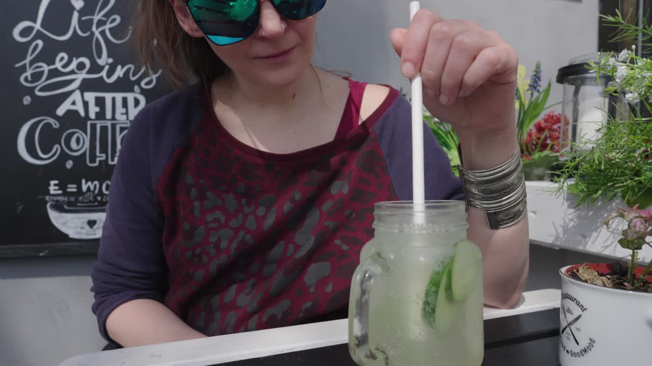 A woman in stylish black sunglasses stirs her cucumber lemonade with a straw at an outdoor restaurant