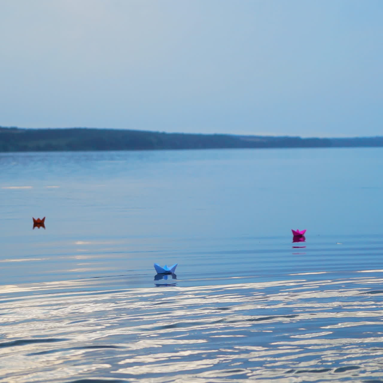 Colored paper ships go far away from the shore on the blue water background at sunset. Homemade boats with different colors floating on the big pond
