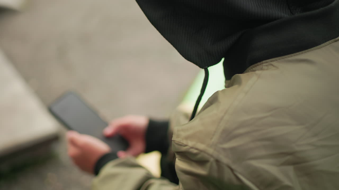 Rear close up of kid in hooded jacket holding smartphone with both hands outdoors, deeply engaged in screen activity, showing focused body posture and natural casual environment around