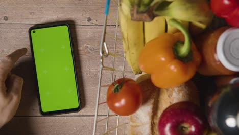 overhead studio shot of person using green screen mobile phone next to basic food items in supermarket wire shopping basket 3