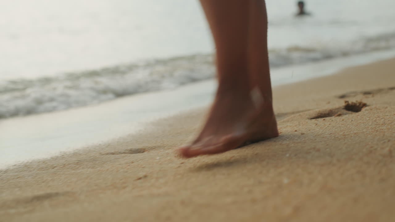 Piedi che camminano sulla spiaggia, giovane donna che passeggia lungo la riva all'ora d'oro con dolci onde che lambiscono, sabbia calda che imprime impronte, atmosfera estiva rilassata, sensazione di viaggio e consapevolezza.