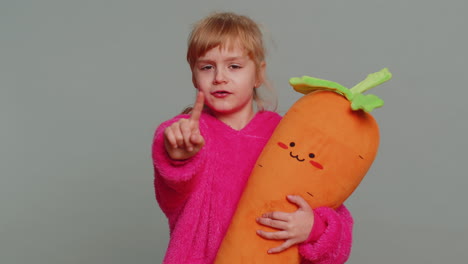 a young girl in pink pajamas holds a carrot plush toy and points at the camera