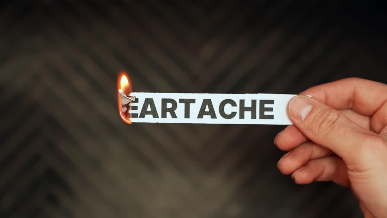 Hand holds a paper strip reading “HEARTACHE” as the edge burns in flame, symbolizing emotional pain, breakup, grief, and relationship loss concept footage